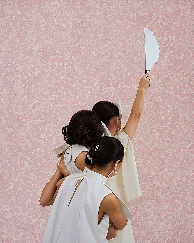 Three women in white sleeveless tops holding a curved blade against pink floral wallpaper